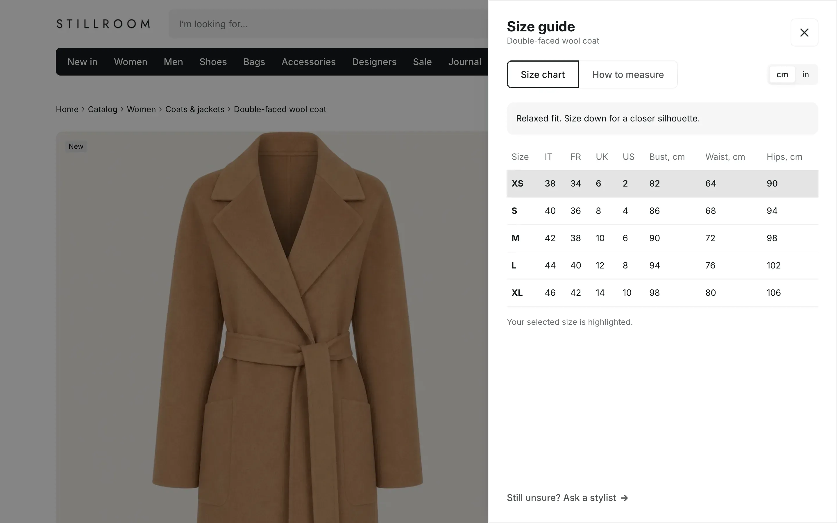 Size guide panel over the product page: the product name, Size chart and How to measure tabs, a cm and in switch, a fit note, a size table with the XS row highlighted, and a Still unsure? Ask a stylist link at the bottom.